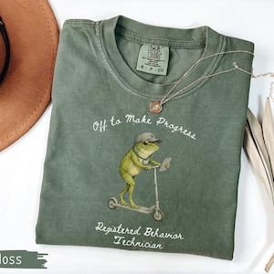 May include: Moss green t-shirt with a frog graphic riding a scooter, wearing a cap and glasses, and holding a tablet. The shirt has the text "Off to Make Progress" and "Registered Behavior Technician". A brown hat and necklace are also visible.