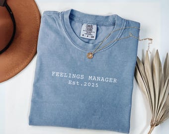 May include: A light blue t-shirt with the words "FEELINGS MANAGER Est. 2025" printed in white. A gold necklace with a pendant rests on the shirt. A brown hat and dried plant are in the background.