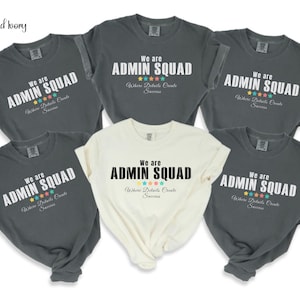 May include: A collection of gray and cream-colored t-shirts. Each shirt displays the text "We are ADMIN SQUAD" in bold letters, with the phrase "Where Details Create Success" below. Colorful stars accent the design.
