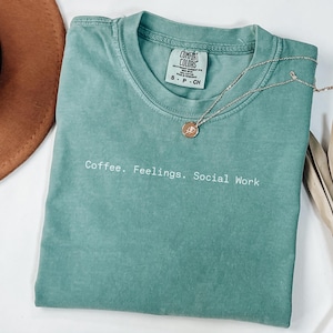 May include: A seafoam green Comfort Colors t-shirt with the text "Coffee. Feelings. Social Work" in white. A gold pendant necklace and a brown hat are also in the image. A dried palm leaf is on the right.