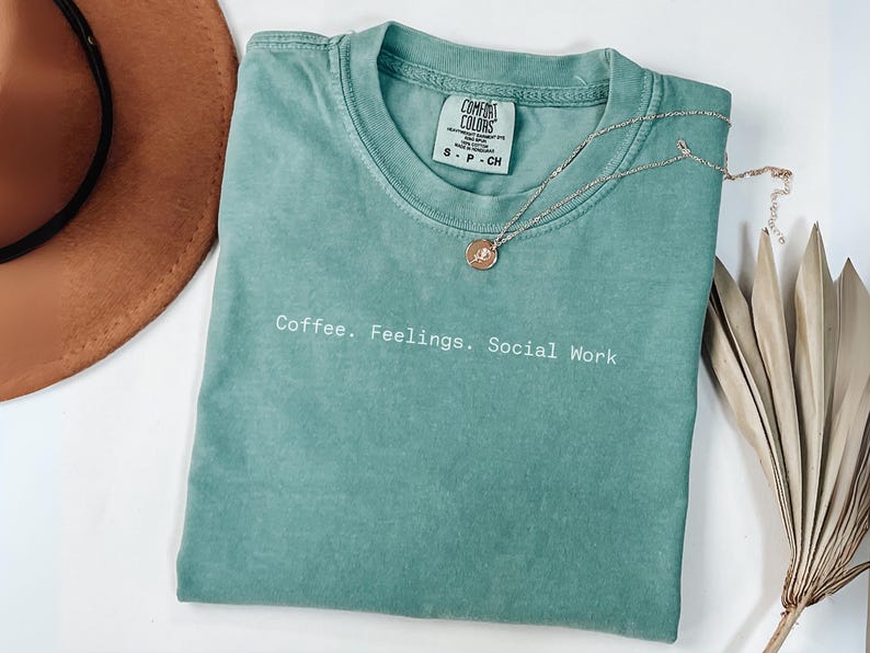 May include: A seafoam green Comfort Colors t-shirt with the text "Coffee. Feelings. Social Work" in white. A gold pendant necklace and a brown hat are also in the image. A dried palm leaf is on the right.