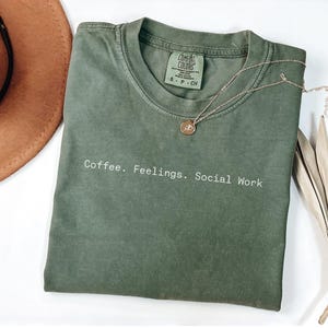 May include: An olive green t-shirt with the words "Coffee. Feelings. Social Work" printed in white. A gold necklace with a pendant and a brown hat are also in the image.