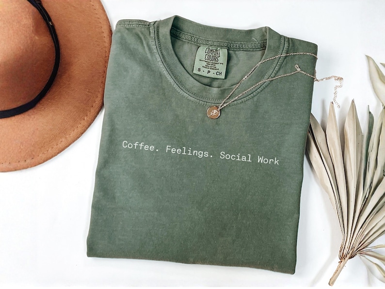 May include: An olive green t-shirt with the words "Coffee. Feelings. Social Work" printed in white. A gold necklace with a pendant and a brown hat are also in the image.