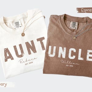 May include: Two t-shirts, one ivory and one espresso, with the words "AUNT" and "UNCLE" printed in distressed brown and white, respectively. Each shirt includes a name and "EST. 2026". Both shirts have a Comfort Colors tag and a gold necklace.