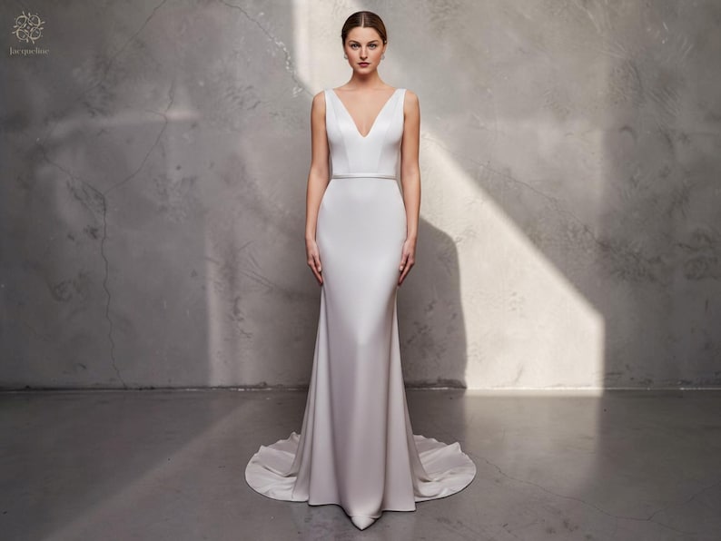 May include: A white bridal gown with a V-neck and a fitted shape. The dress has a slight train and a thin belt at the waist. The model is standing in front of a grey textured wall.