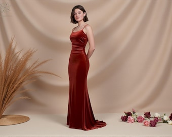 May include: A floor-length, rust-colored velvet gown with spaghetti straps. The dress features a fitted bodice and a flowing skirt with a slight train. The model is standing with hands behind the back. A bouquet of flowers is on the floor.