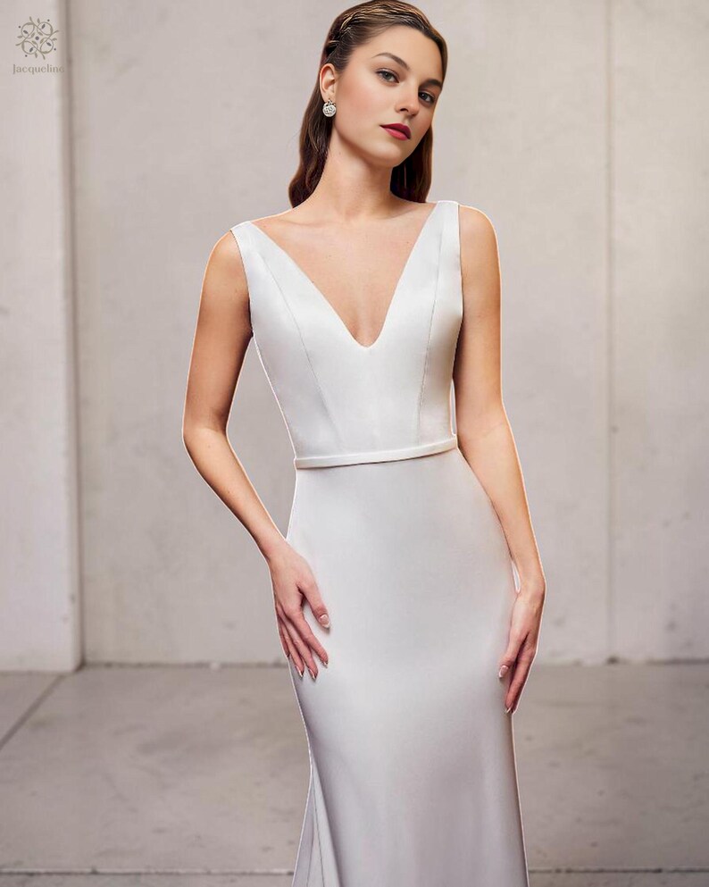 May include: A white wedding dress with a plunging V-neck and a fitted shape. The sleeveless gown has a thin belt at the waist and a slight train. The model wears earrings and has her hair styled back.