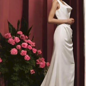 May include: A woman wearing a white sleeveless gown with a cowl neck and a long train. The gown is made of a smooth, satin-like fabric. The woman is standing in front of a pink wall with a bouquet of pink roses in the background.