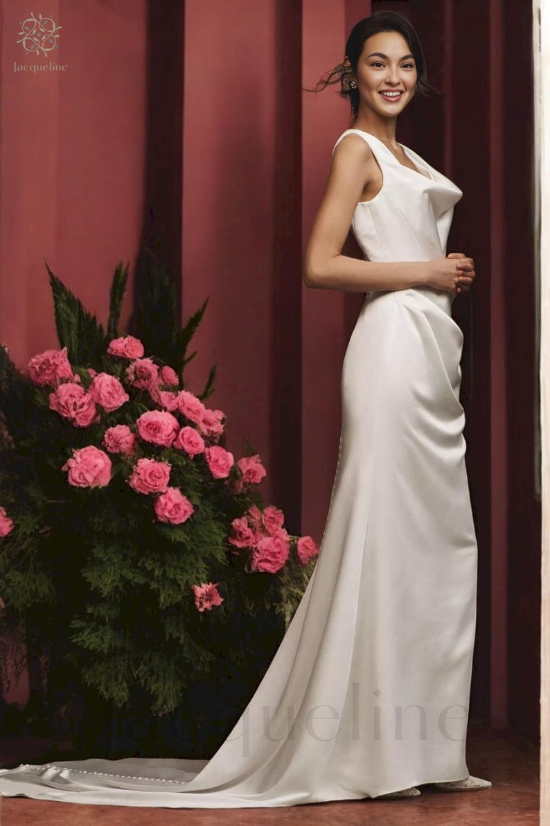 May include: A woman wearing a white sleeveless gown with a cowl neck and a long train. The gown is made of a smooth, satin-like fabric. The woman is standing in front of a pink wall with a bouquet of pink roses in the background.