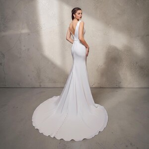 May include: A white bridal gown with a mermaid silhouette and a long train. The dress has a V-neck, a fitted bodice, and buttons down the back. The model is standing in front of a grey wall.