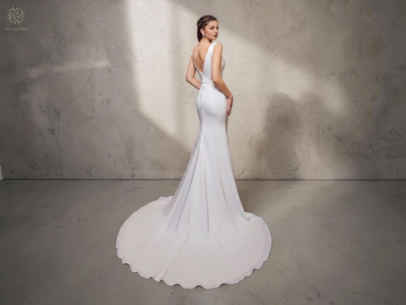 May include: A white bridal gown with a mermaid silhouette and a long train. The dress has a V-neck, a fitted bodice, and buttons down the back. The model is standing in front of a grey wall.