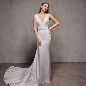 May include: A white, sleeveless wedding dress with a V-neck and a flowing train. The gown features a fitted bodice and a delicate belt at the waist. The fabric has a smooth, satin-like appearance. The model is standing in front of a grey wall.