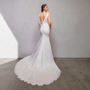 May include: A white bridal gown featuring a V-neck and a long, trailing train. The dress has a fitted bodice and a buttoned back. The skirt gently flares from the knees. The fabric appears soft and flowing.