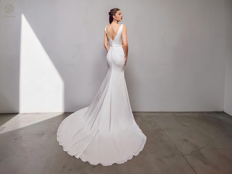 May include: A white bridal gown featuring a V-neck and a long, trailing train. The dress has a fitted bodice and a buttoned back. The skirt gently flares from the knees. The fabric appears soft and flowing.