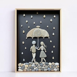 Peut inclure: Une œuvre d'art encadrée représentant un couple sous un parapluie, réalisée avec des galets. Le parapluie et les personnages sont faits de pierres grises, sur fond sombre avec des gouttes de pluie en galets. Le cadre est doré.