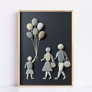 May include: A framed artwork featuring a family made of stones on a black background. The child holds stone balloons. The parents are holding hands and carrying purses. The frame is gold.