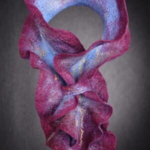 May include: A felted scarf featuring a deep magenta exterior and a light blue interior. The fabric has a wavy, ruffled texture with subtle gold-colored fibers woven throughout the material, creating a layered, sculptural appearance with soft edges.