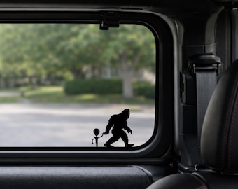 May include: Black vinyl decal featuring a silhouette of Bigfoot walking and holding hands with a small alien. The decal is applied to a vehicle window, with a blurred outdoor scene visible in the background.