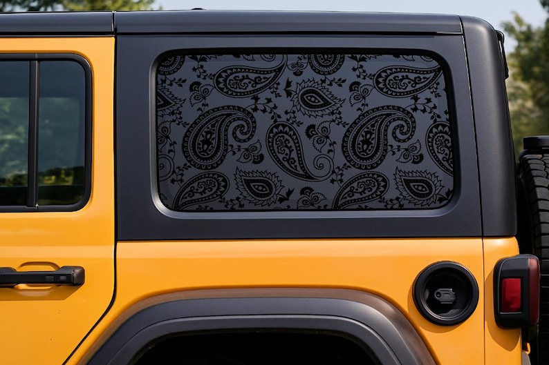 May include: A close-up of a yellow Jeep with a black window featuring a black paisley pattern. The Jeep's rear window is decorated with a repeating design of black paisley shapes. The vehicle's black trim and a spare tire are also visible.