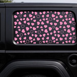 May include: Black vehicle window with a pink heart pattern. The hearts are in various sizes and styles, some solid and some outlined, against a dark background. The design is repeated across the window.