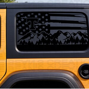 Può includere: Un adesivo in vinile nero applicato sul finestrino laterale posteriore di un veicolo giallo. Il design presenta una bandiera americana stilizzata con una catena montuosa e una silhouette di foresta, con una grande lettera T in alto a sinistra.