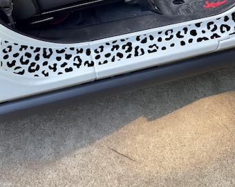 May include: White vehicle door sill protector with black leopard print pattern. The protective strip is installed on the lower edge of the door frame. The vehicle's interior is visible in the background.