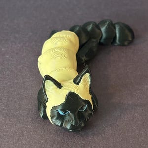May include: A 3D printed figurine of a Siamese cat curled up in a sleeping position. The cat is cream colored with black markings and blue eyes.