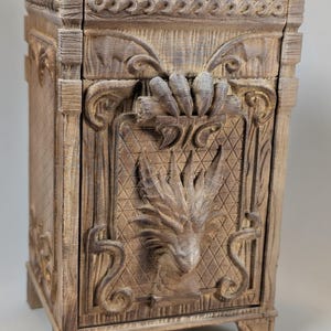 May include: A wooden cabinet with intricate carvings. The cabinet features a detailed dragon head and floral designs. The color is a light brown with darker shading, giving it a rustic appearance. The cabinet has a rectangular shape and stands on four small feet.