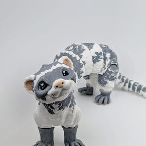 May include: A handcrafted, whimsical sculpture of a ferret. The ferret is primarily white with gray markings and a gray tail. The sculpture has a friendly expression and is posed on all fours. The background is a plain white.