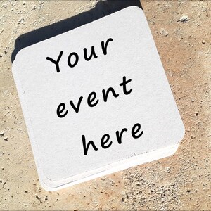 May include: A stack of white square coasters with rounded corners. The text "Your event here" is printed in black on the top coaster. The coasters are on a textured, light brown surface.