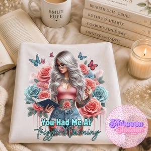 May include: White t-shirt featuring a graphic of a woman with tattoos, glasses, and long gray hair, surrounded by roses and butterflies. Text on the shirt reads "You Had Me At Trigger Warning."