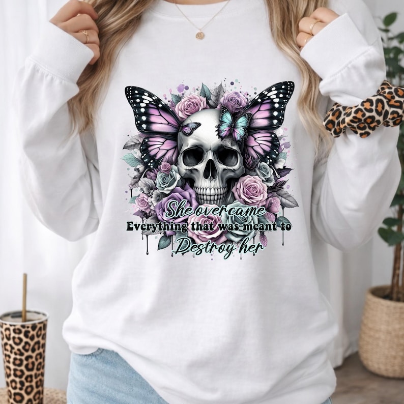 May include: White long-sleeve shirt featuring a skull with butterfly wings and floral accents in shades of purple, pink, and blue. The text "She overcame everything that was meant to destroy her" is also visible.