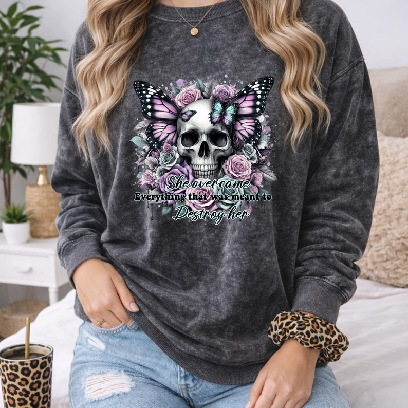 May include: Dark gray sweatshirt featuring a skull design with butterfly wings and floral accents in pink, purple, and blue. The text "She overcame Everything that was meant to Destroy her" is printed below the skull.