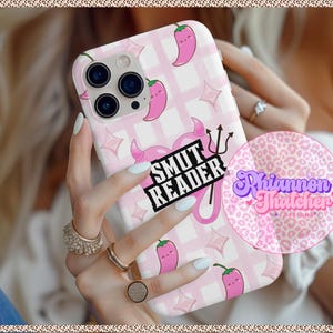 May include: A pink and white phone case featuring a repeating pattern of pink chili peppers and stars. The case has the text "SMUT READER" and a devil graphic. The phone case is held by a person with multiple rings.