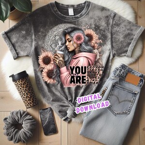 May include: A grey t-shirt featuring a graphic of a woman with grey hair, sunflowers, and the words "YOU ARE" with inspirational words. The image includes a leopard print travel mug, a scrunchie, a phone, and jeans.
