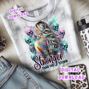 May include: White t-shirt with a graphic of a woman with colorful hair and a back tattoo, surrounded by skulls. The text "YOU ARE STRONGER THAN THE STORM" is featured. The words "DIGITAL DOWNLOAD" are also visible.