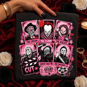 May include: A black t-shirt featuring a graphic print of six horror movie characters in pink-bordered frames. The characters include Freddy Krueger, Pennywise, Chucky, Jason Voorhees, Ghost Face with a censored mask, and Michael Myers. Text includes THE DREAM, THE CLOWN, THE KILLER, GHOST FACE, THE BUTCHER, CUT, and SPOOKY MOVIE NIGHT.