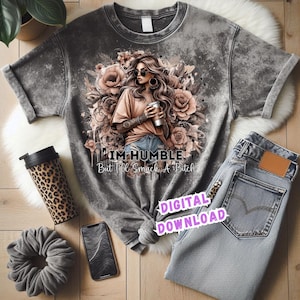 Op de afbeelding: Een grijs t-shirt in tie-dye stijl met een afbeelding van een vrouw die een koffiekopje vasthoudt, omringd door bloemen. De tekst "I'M HUMBLE But I'll Smack A Bitch" staat eronder. Een spijkerbroek, een reismok met luipaardprint, een scrunchie en een telefoon staan ook in de afbeelding.