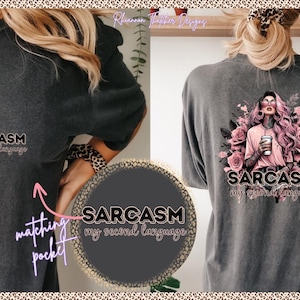 May include: Dark gray t-shirt with the text "Sarcasm my second language." The back of the shirt features a graphic of a woman with pink hair, sunglasses, and a coffee cup, surrounded by pink roses. The shirt has a matching pocket.