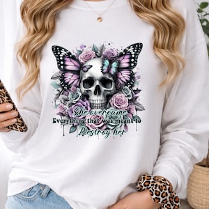 May include: White long-sleeved shirt featuring a skull design with butterfly wings and floral accents in shades of purple, pink, and teal. The text "She overcame everything that was meant to destroy her" is printed below the skull.