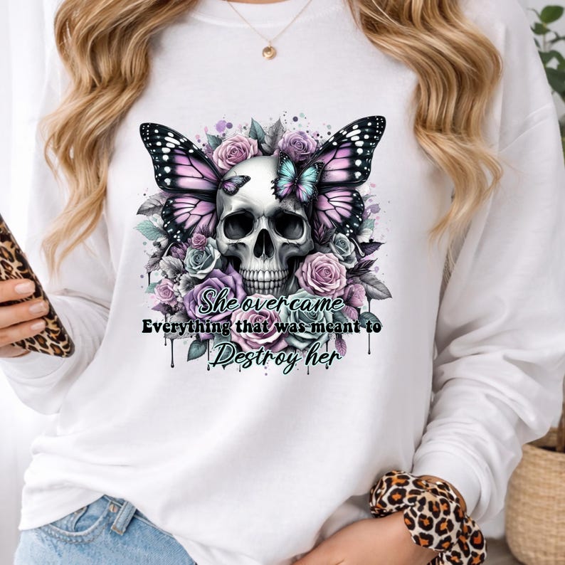 May include: White long-sleeved shirt featuring a skull design with butterfly wings and floral accents in shades of purple, pink, and teal. The text "She overcame everything that was meant to destroy her" is printed below the skull.