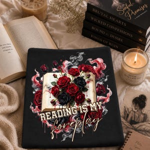 May include: Black graphic tee with a floral design of red and black roses, an open book, and the text "READING IS MY foreplay." The image includes books, a candle, and a cup of coffee.
