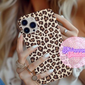 May include: A phone case with a leopard print design. The case features a tan background with brown spots. The phone case is held in a person's hands. The phone case has a camera bump with three lenses.