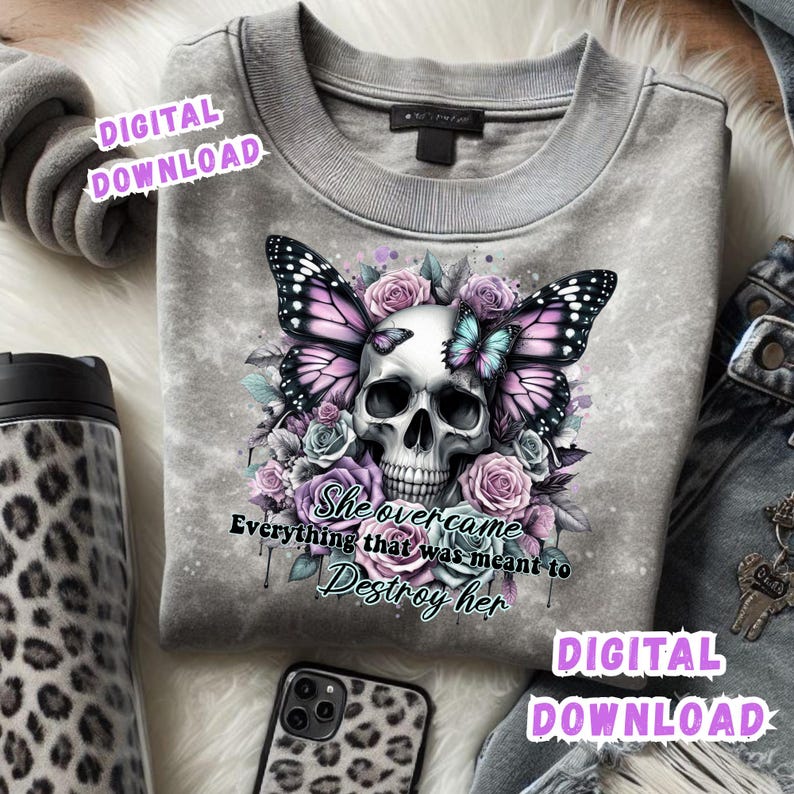 May include: A gray sweatshirt featuring a skull with butterfly wings and floral accents in shades of purple, pink, and blue. The text "She overcame everything that was meant to destroy her" is below the skull. A leopard print phone case and tumbler are visible.