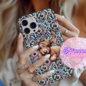 May include: A phone case with a floral and butterfly design, featuring a Highland cow illustration. The case has a turquoise, purple, and brown colour scheme. The case is held by a person with multiple rings on their fingers.