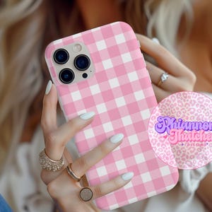 Può includere: Custodia per telefono con motivo a quadretti rosa e bianchi. La custodia ha un motivo quadrato e un ritaglio per l'obiettivo della fotocamera. È visibile un adesivo circolare con la scritta "Rhiannon Thatcher Designs".