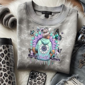Puede incluir: Sudadera gris con un gráfico de una mujer meditando, rodeada de mariposas y flores. El texto "Mental Health Matters" está debajo. La sudadera está doblada sobre una superficie blanca, con un vaso y una funda de teléfono con estampado de leopardo.