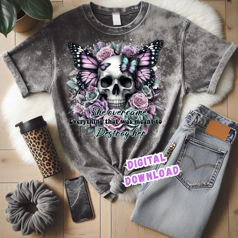 May include: Gray t-shirt with a skull and butterfly graphic, surrounded by roses. The text reads "She overcame everything that was meant to destroy her." Also pictured are jeans, a leopard-print tumbler, a scrunchie, and a smartphone.