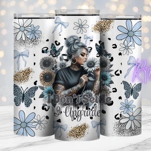 May include: A white tumbler featuring a woman with tattoos, sunflowers, and butterflies. The design includes blue flowers, bows, and leopard print accents. The text "I Don't Settle, I Upgrade" is displayed on the tumbler.