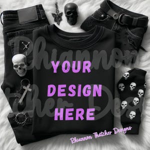 May include: A black crew neck sweatshirt with the text "YOUR DESIGN HERE" in purple, alongside black jeans, a skull-printed sock, and skull-themed accessories. The items are arranged on a white, fluffy surface.